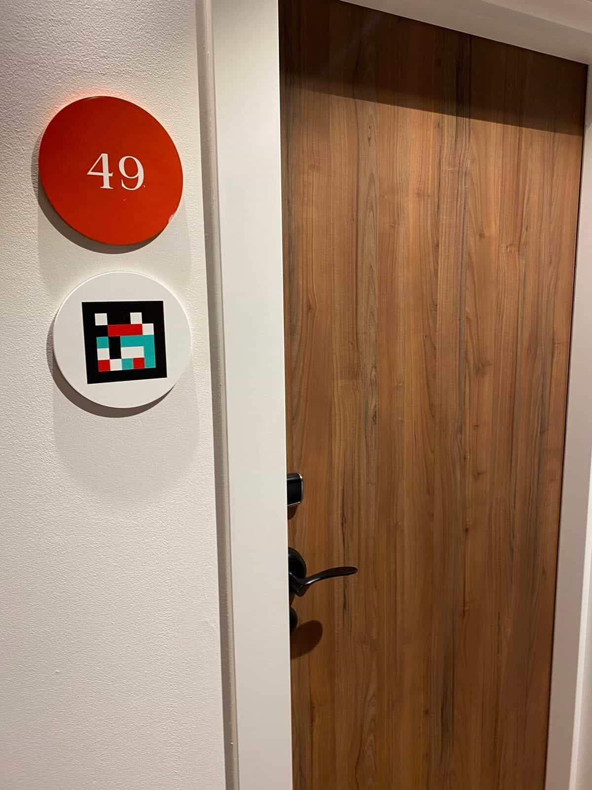 Zimmer 49 am ibis Styles Sevilla: weißer Türrahmen mit orangem Disc und „49“ und direkt darunter NaviLens-Code auf weißem Disc