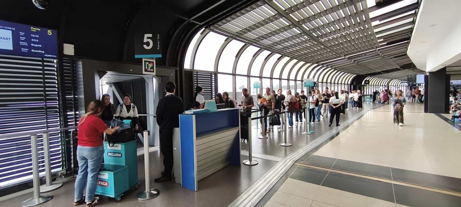 Korridor von Gate 5 am Flughafen José María Córdova mit tonnenförmigem Metallrippendach: schwarzes Hängeschild «5 Puerta · Gate» mit NaviLens-Code darunter, blaues FIDS links mit «JetSMART · Flug 5113 · Ziel Bogotá · 13:13 · Boarding»; am Fuß des Schildes Tresen und Trolley «JetSMART INCLUIDO / SE PAGA» mit wartenden Passagieren