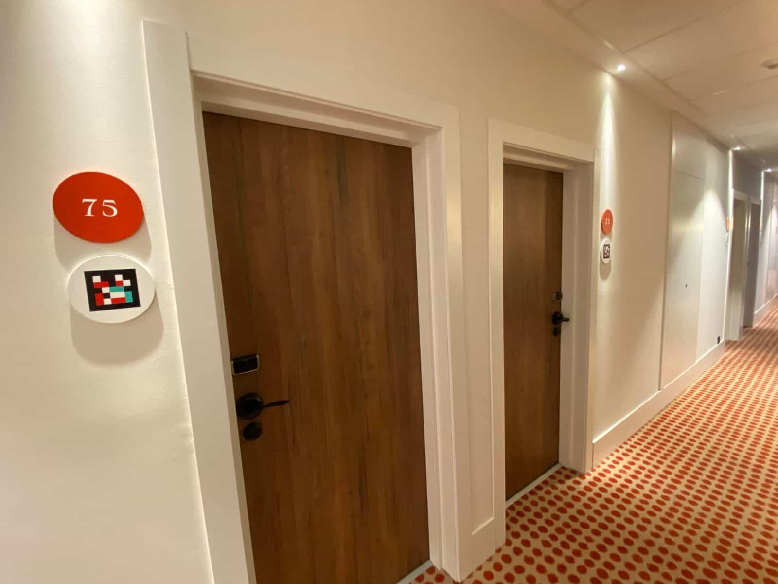 Zimmer 78 am ibis Styles Sevilla: Holztür mit schwarzer Klinke und am weißen Türrahmen ein roter Disc mit „78“ und einem NaviLens-Disc darunter