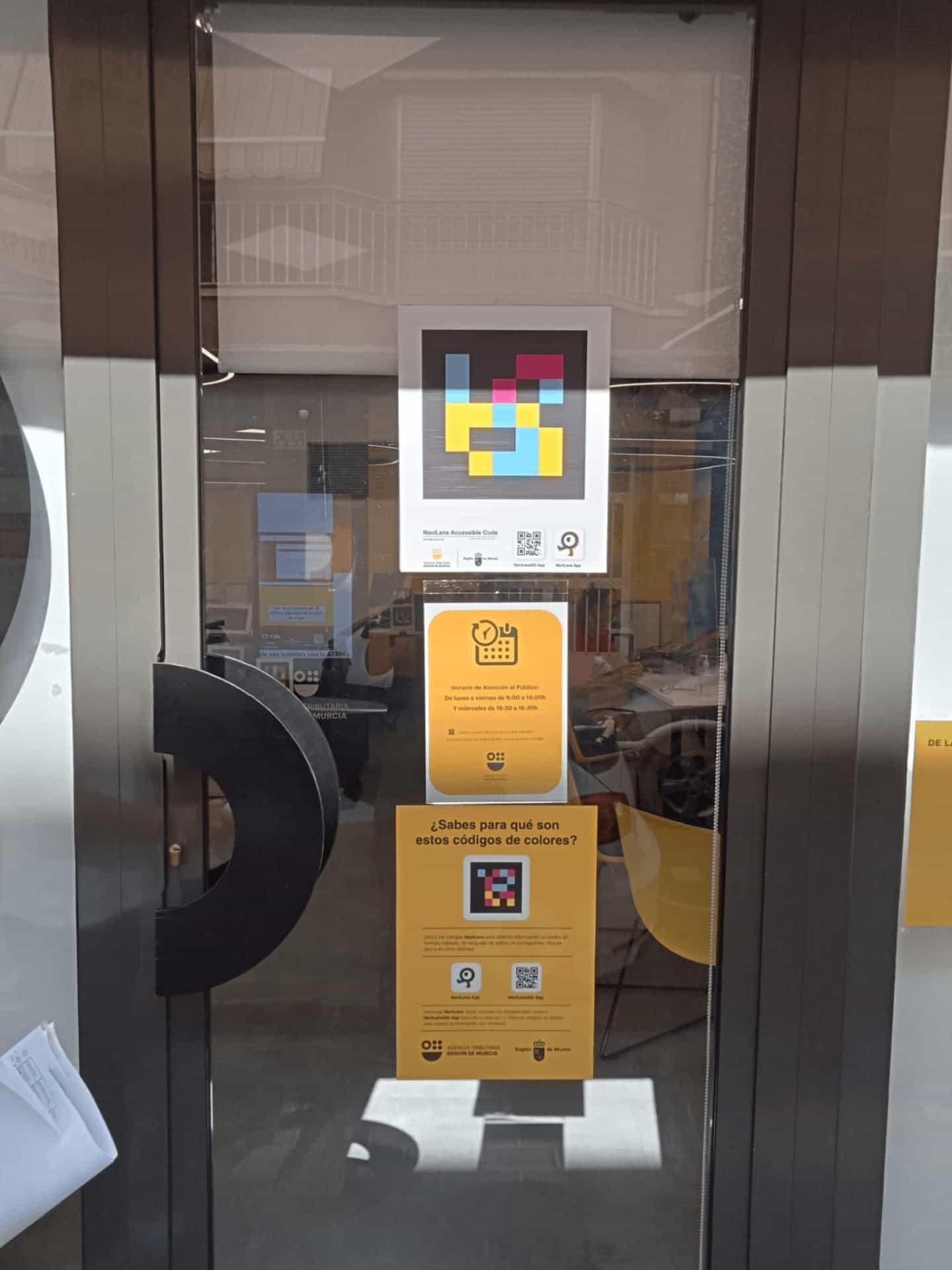 Porta a vetri d’ingresso all’ufficio ATRM di Águilas vista dalla strada, con codice NaviLens, cartello giallo degli orari e volantino esplicativo sui codici a colori.