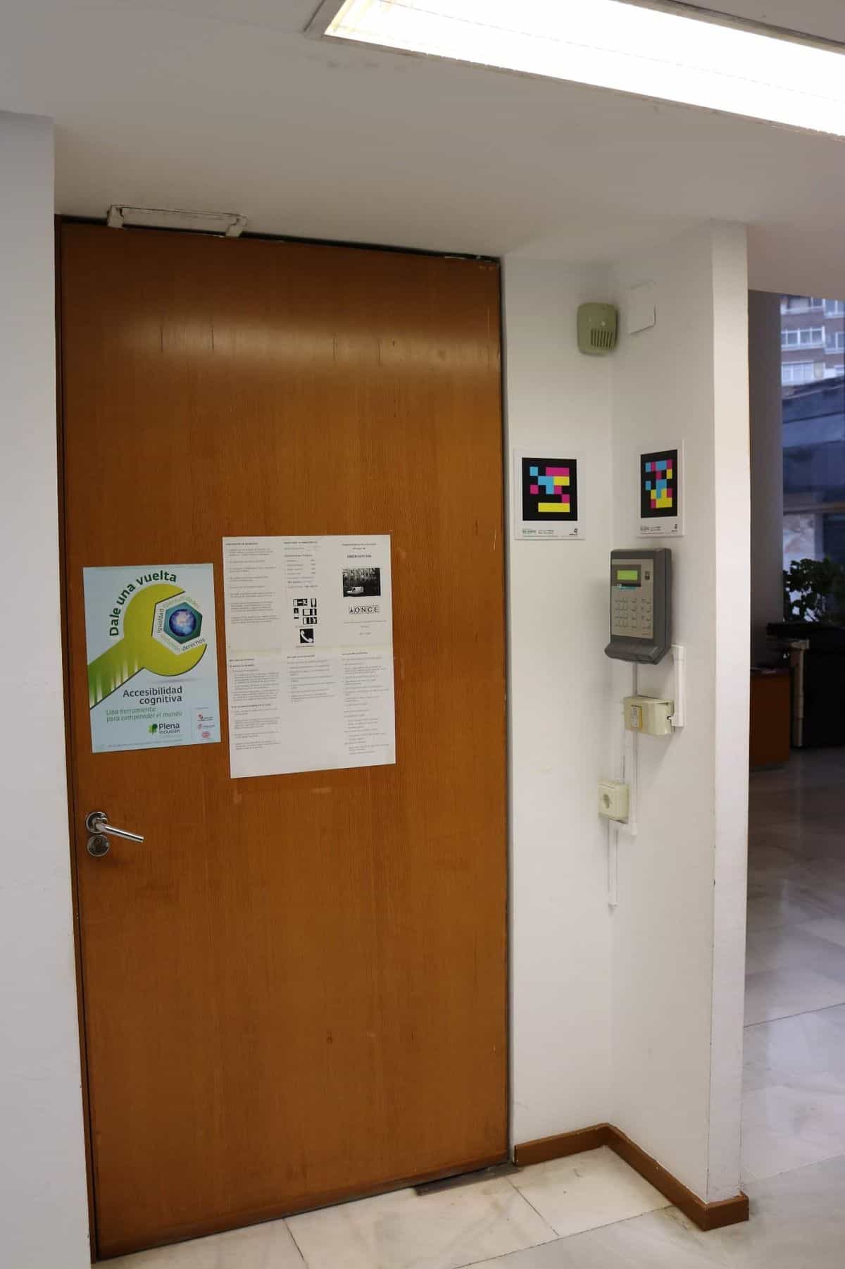 Puerta de madera del centro de control de la ONCE Valladolid con cartel del plan de evacuación y de accesibilidad cognitiva «Dale una vuelta»; a la derecha, dos códigos NaviLens cuadrados a colores junto a un teclado de control de acceso y un dispositivo de detección