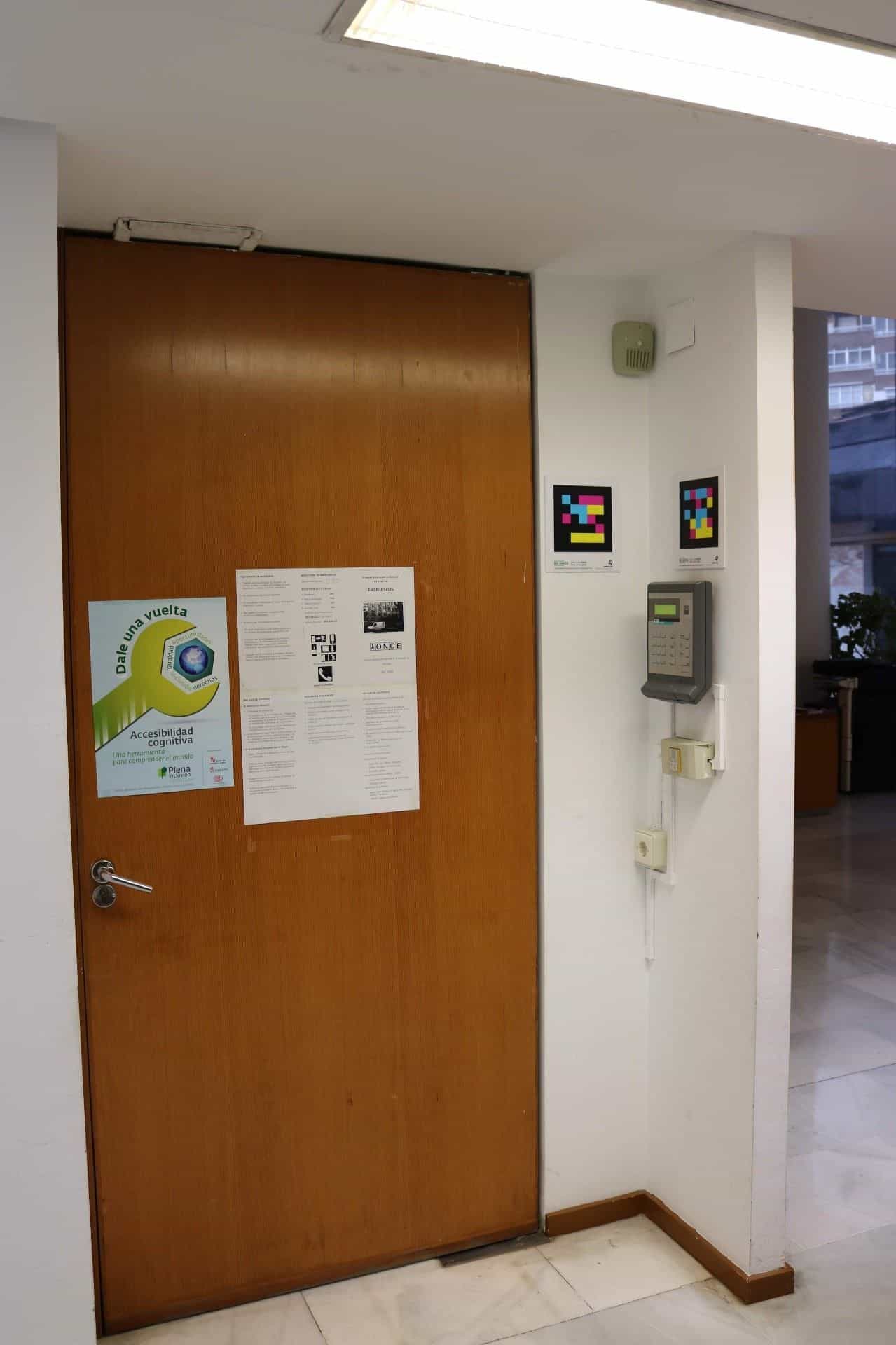 Porta di legno del centro di controllo dell’ONCE Valladolid con il cartello del piano di evacuazione e di accessibilità cognitiva «Dale una vuelta»; a destra, due codici NaviLens quadrati a colori accanto a una tastiera di controllo accessi e a un dispositivo di rilevamento