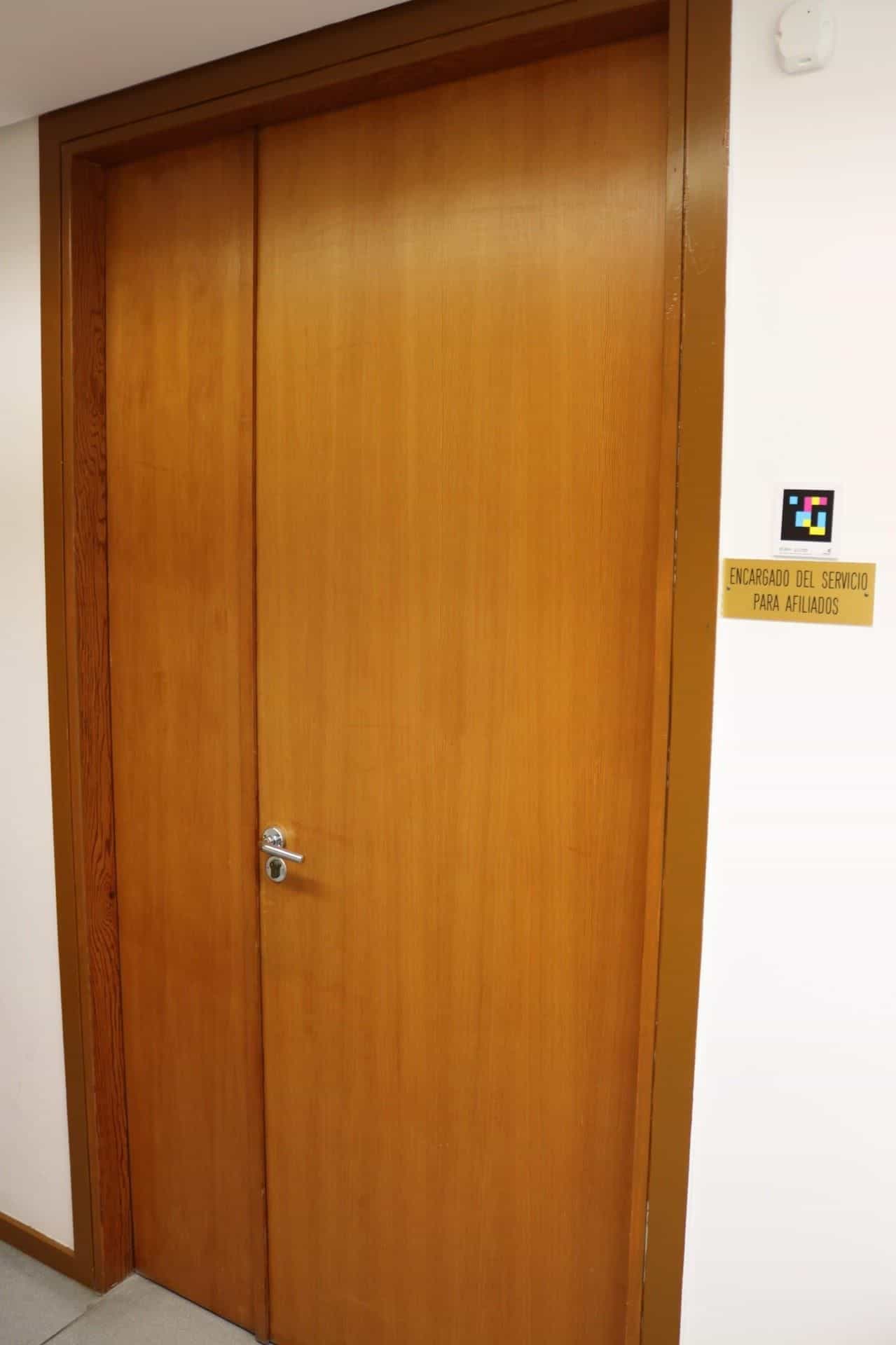 Porta di legno a doppio battente nell’ONCE Valladolid; a destra, una piccola targa dorata con la scritta «ENCARGADO DEL SERVICIO PARA AFILIADOS» e, sopra di essa, un codice NaviLens quadrato a colori in formato piccolo