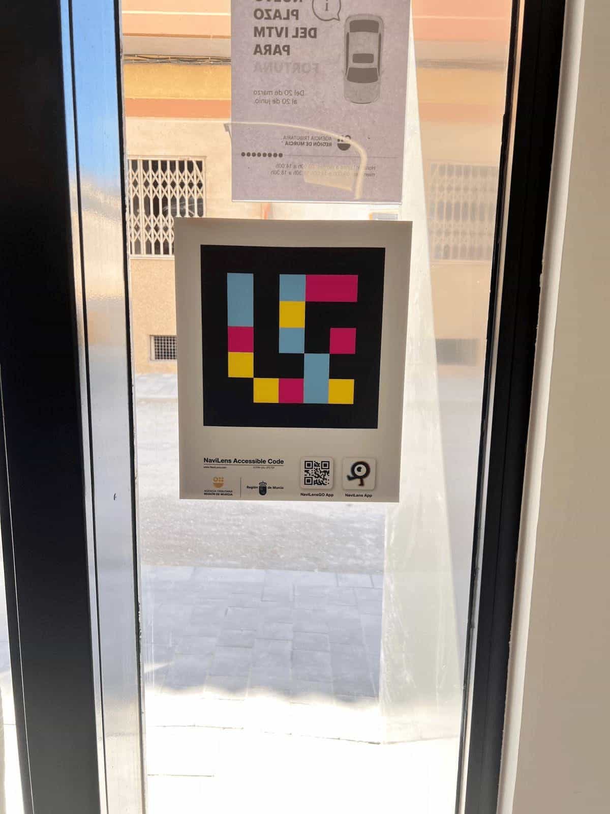 Puerta acristalada de entrada a la oficina ATRM de Fortuna desde el interior, con cartel del nuevo plazo del IVTM y panel del código NaviLens junto al logotipo institucional.