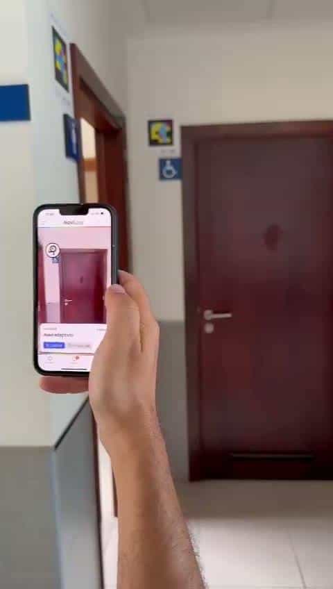 Burgundy door of an accessible toilet in a health centre, with a wheelchair pictogram and a NaviLens code on the wall; a user points their phone at the code and the app shows "Accessible toilet".