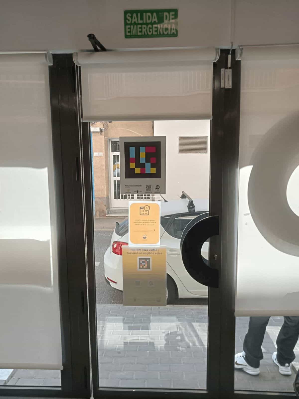 Misma puerta vista desde dentro: cartel verde «SALIDA DE EMERGENCIA», código NaviLens, horario y folleto explicativo espejados, con el logotipo ATRM en vinilo blanco mate.