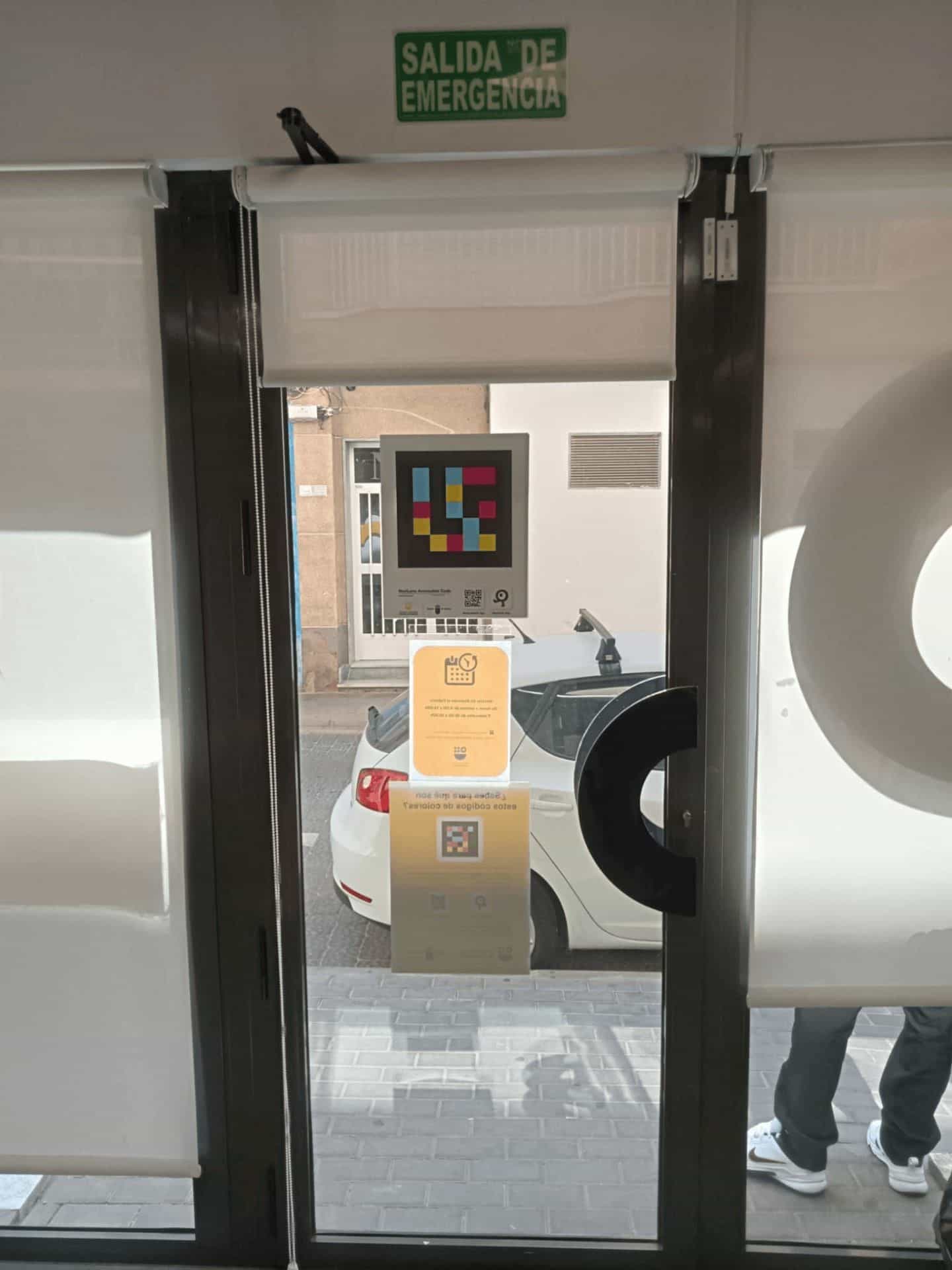 La stessa porta vista da dentro: cartello verde «SALIDA DE EMERGENCIA», codice NaviLens, orario e volantino esplicativo rispecchiati, con il logo ATRM in vinile bianco opaco.