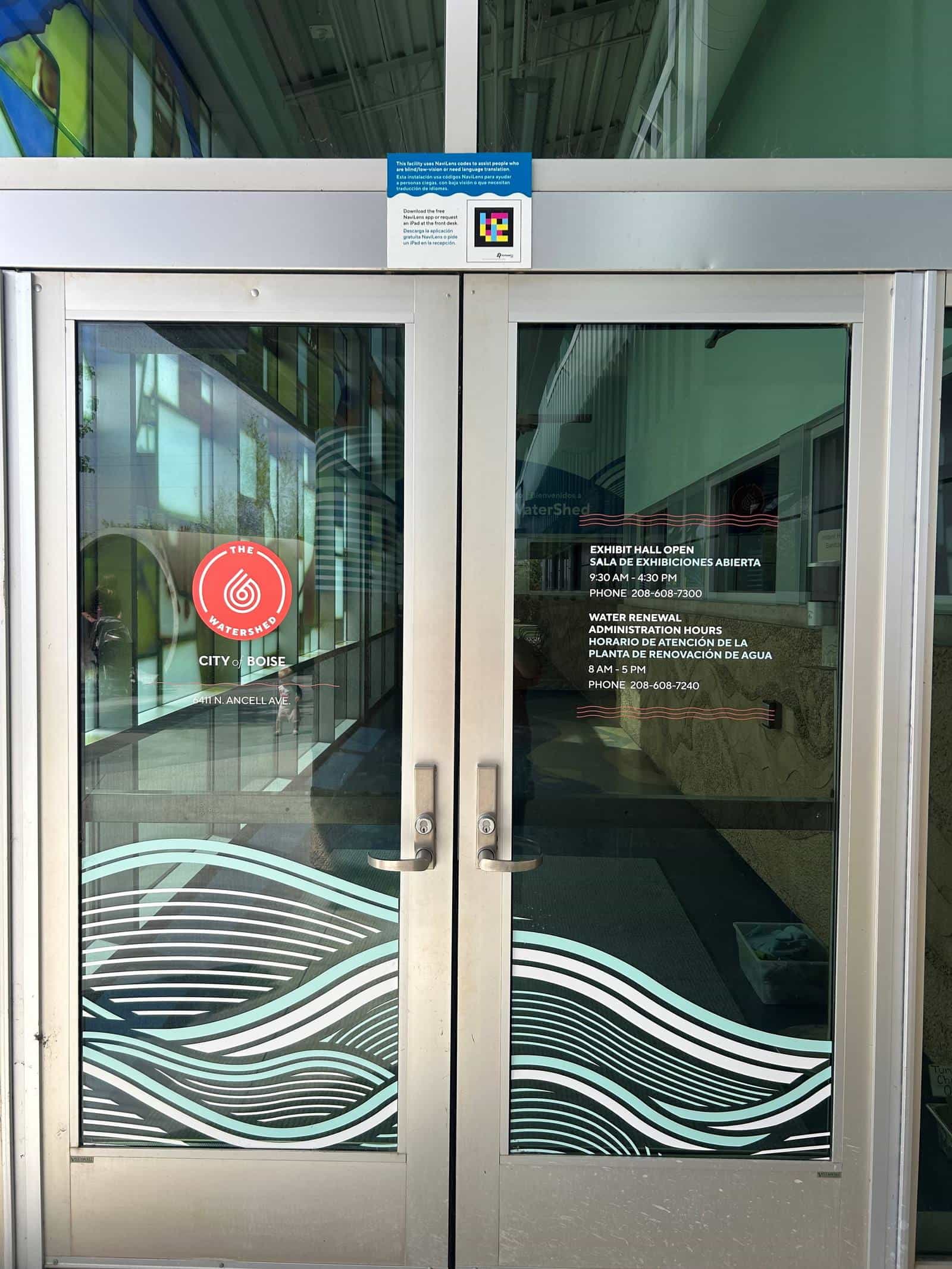 The glass entrance doors at The WaterShed, with the centre's logo, address and hours lettered in English and Spanish; above the transom, a bilingual blue sign announcing the NaviLens codes, with its own code printed beside it