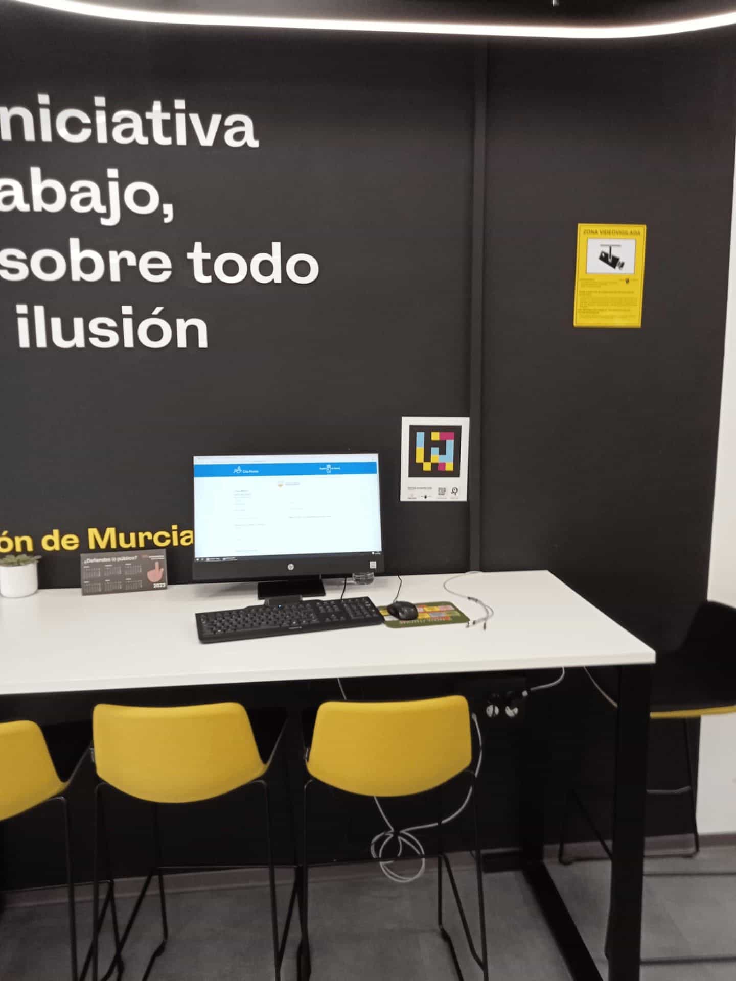 Postazione self-service per la prenotazione con monitor che mostra il sito «Cita Previa Región de Murcia», targa NaviLens sulla parete e murale «iniziativa, lavoro, soprattutto entusiasmo».
