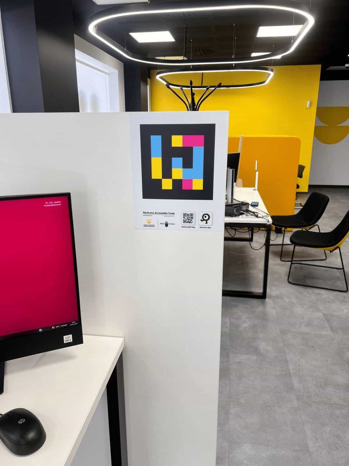 Mampara blanca lateral del puesto interior con código NaviLens a la altura de la vista, monitor HP encendido y sala diáfana «store» con mural amarillo al fondo.
