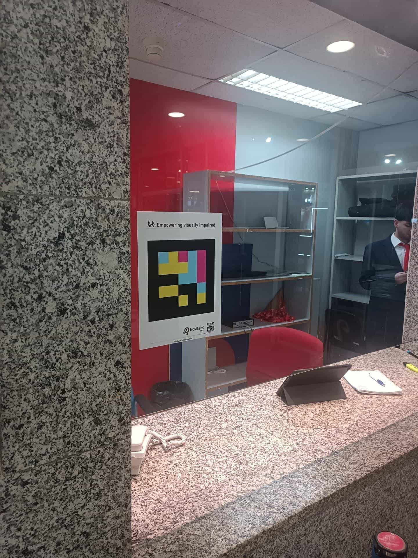 Punto informativo di IFEMA MADRID: sul vetro del banco un cartello «Empowering visually impaired» con un codice NaviLens quadrato di grandi dimensioni e la scritta «Punto informativo»
