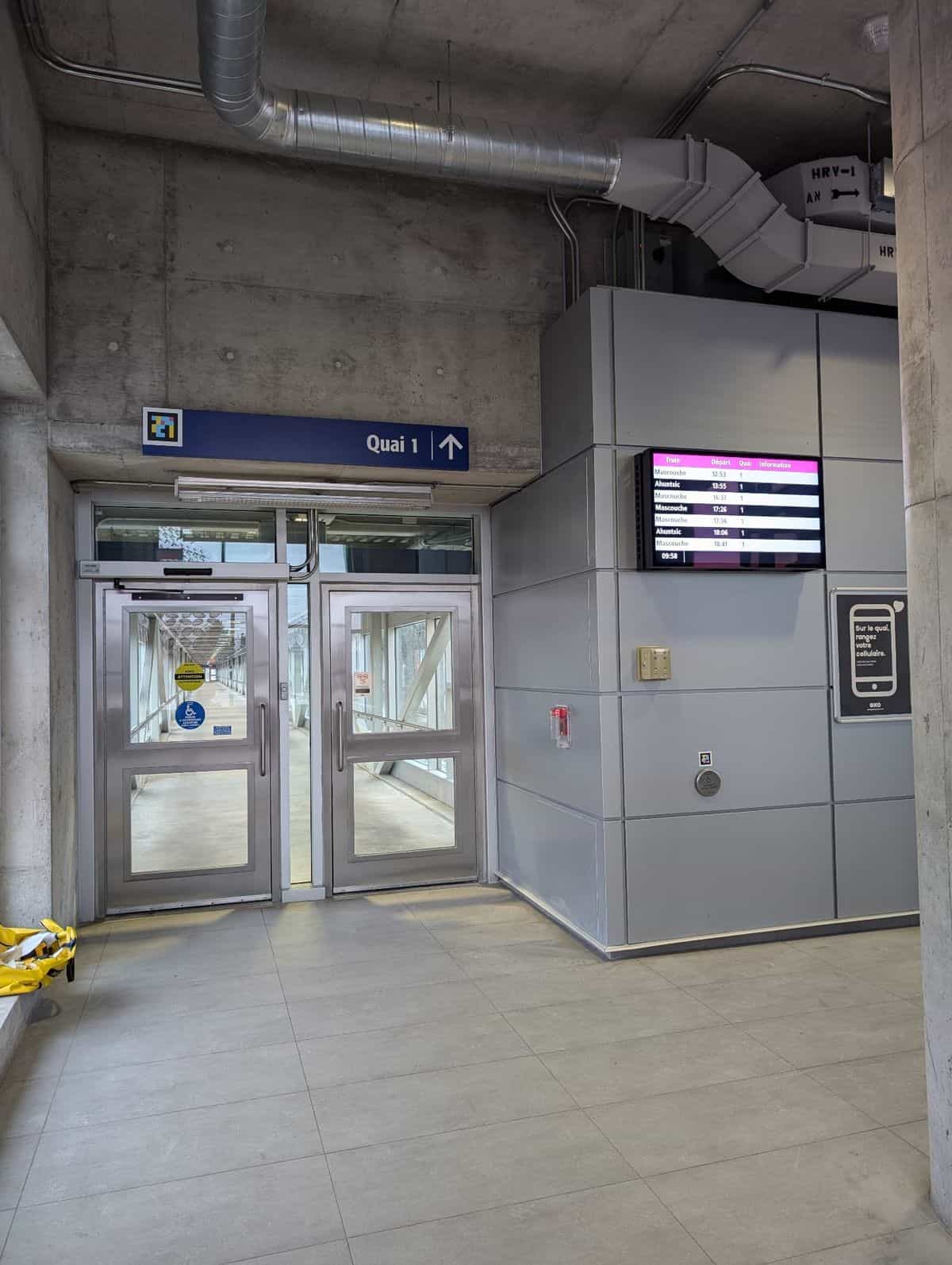 Vestibule of the «Quai 1» walkway at an exo Mascouche-line station: exposed concrete walls and two metal-framed glass doors; on the lintel, exo's blue-and-yellow «Quai 1 ↑» sign with the NaviLens code in cyan, magenta and yellow