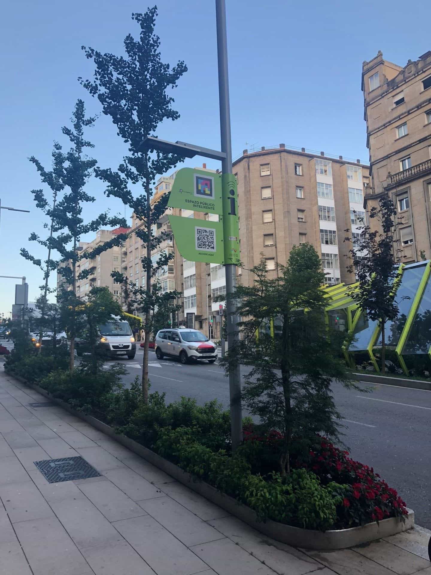 Veduta laterale della nuova Gran Vía di Vigo, con fioriere continue piene di alberi giovani, arbusti e fiori rossi; un palo della luce sostiene una targa verde dell’Espazo Público Intelixente con codice NaviLens GO e QR «Accede aquí» e, in fondo, le coperture verdi delle nuove rampe mobili umanizzate