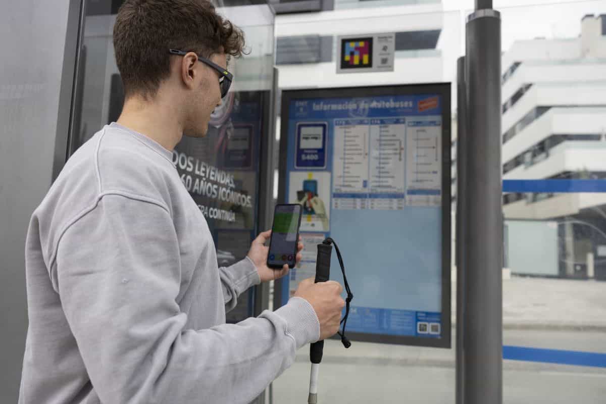 Giovane con bastone bianco che usa l’app NaviLens a una pensilina della EMT a Madrid