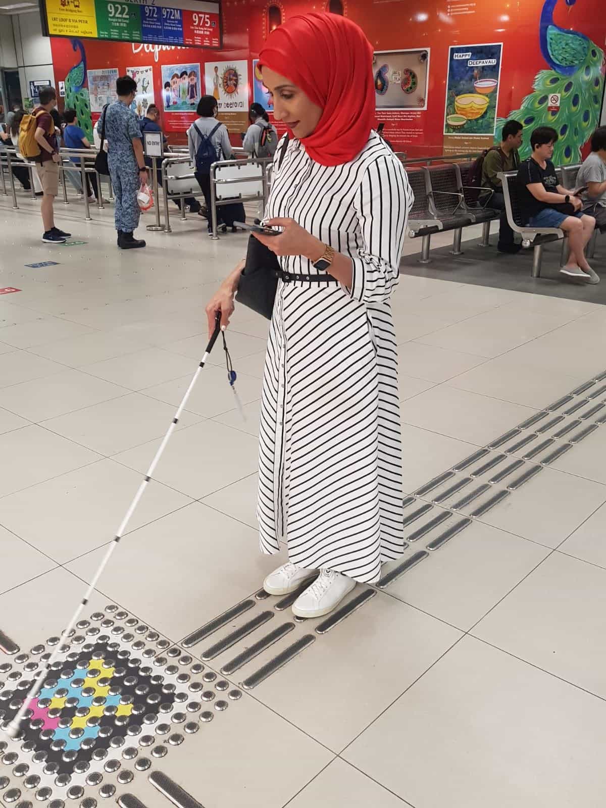 Donna con bastone bianco che cammina in una stazione intermodale di Singapore, seguendo la segnaletica accessibile a terra