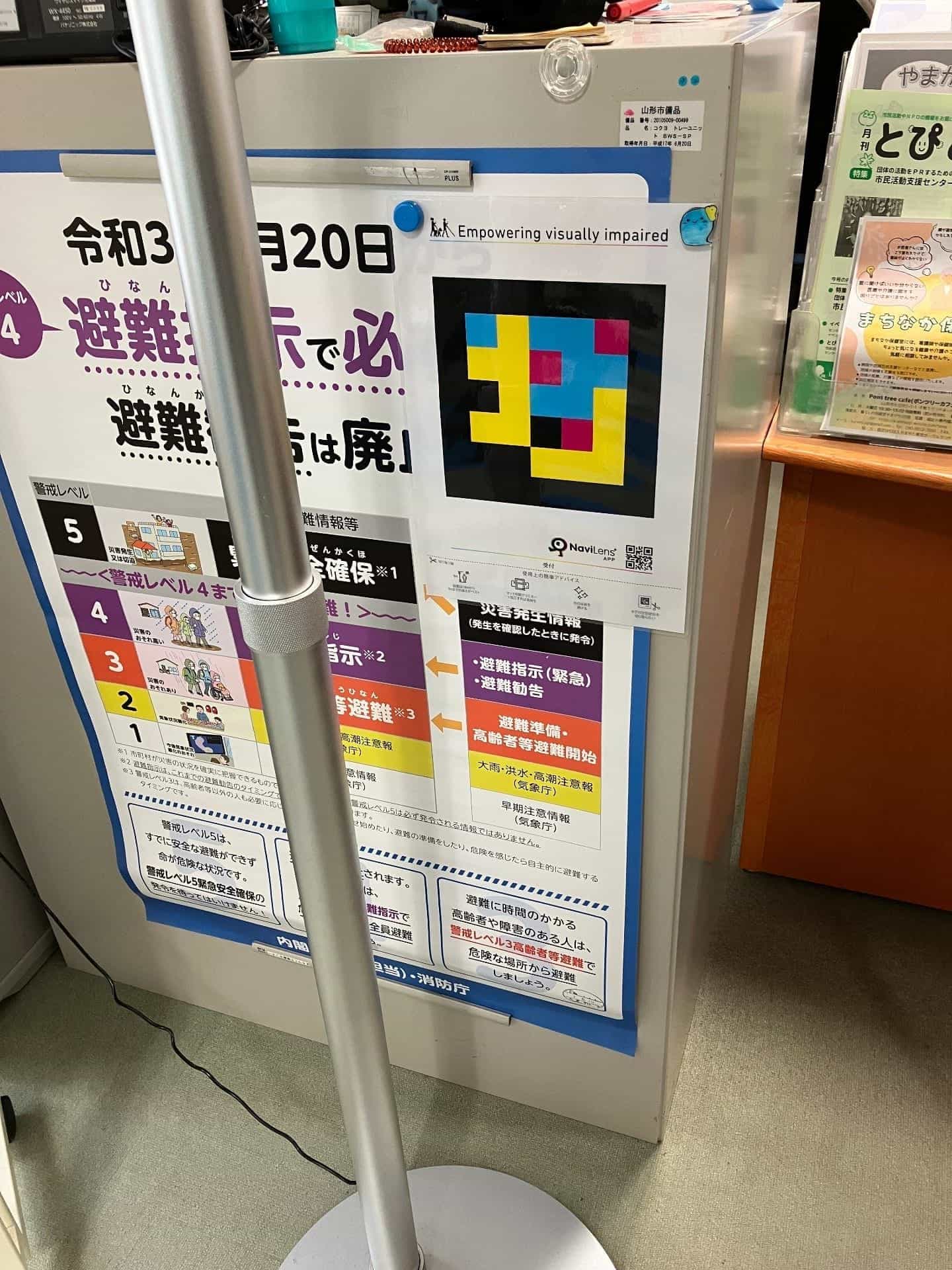 Comptoir d’accueil (受付) du centre civique de Yamagata avec code NaviLens à côté du panneau de niveaux d’alerte d’évacuation 避難指示 et de la revue « とぴあ »