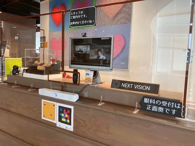 神戸アイセンター2階、NEXT VISION(ビジョンパーク)の受付カウンター:白とコンベンショナルの2つのNaviLensタグが「NEXT VISION」のサイン、スタッフが映るビデオ通話画面、カラフルなハートの壁画の脇に並ぶ