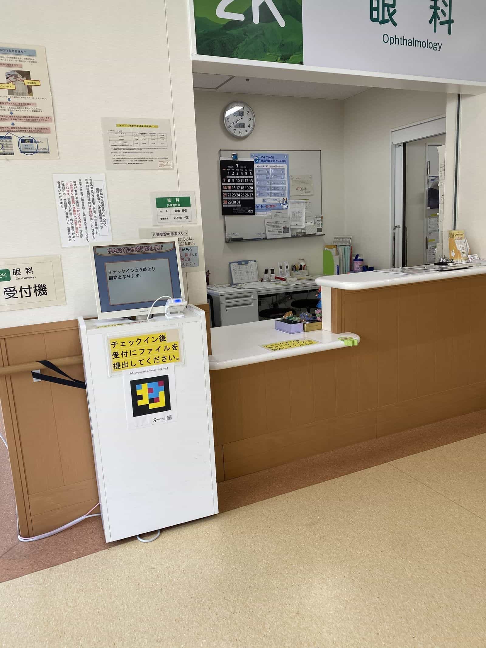 大分大学医学部附属病院 眼科の受付エリア：明るい木目調カウンターの前に白いチェックイン用トーテムが設置され、その前面、黄色の日本語表示の下に「Empowering visually impaired」と書かれたラミネート紙のNaviLensカラータイルコードが貼られている