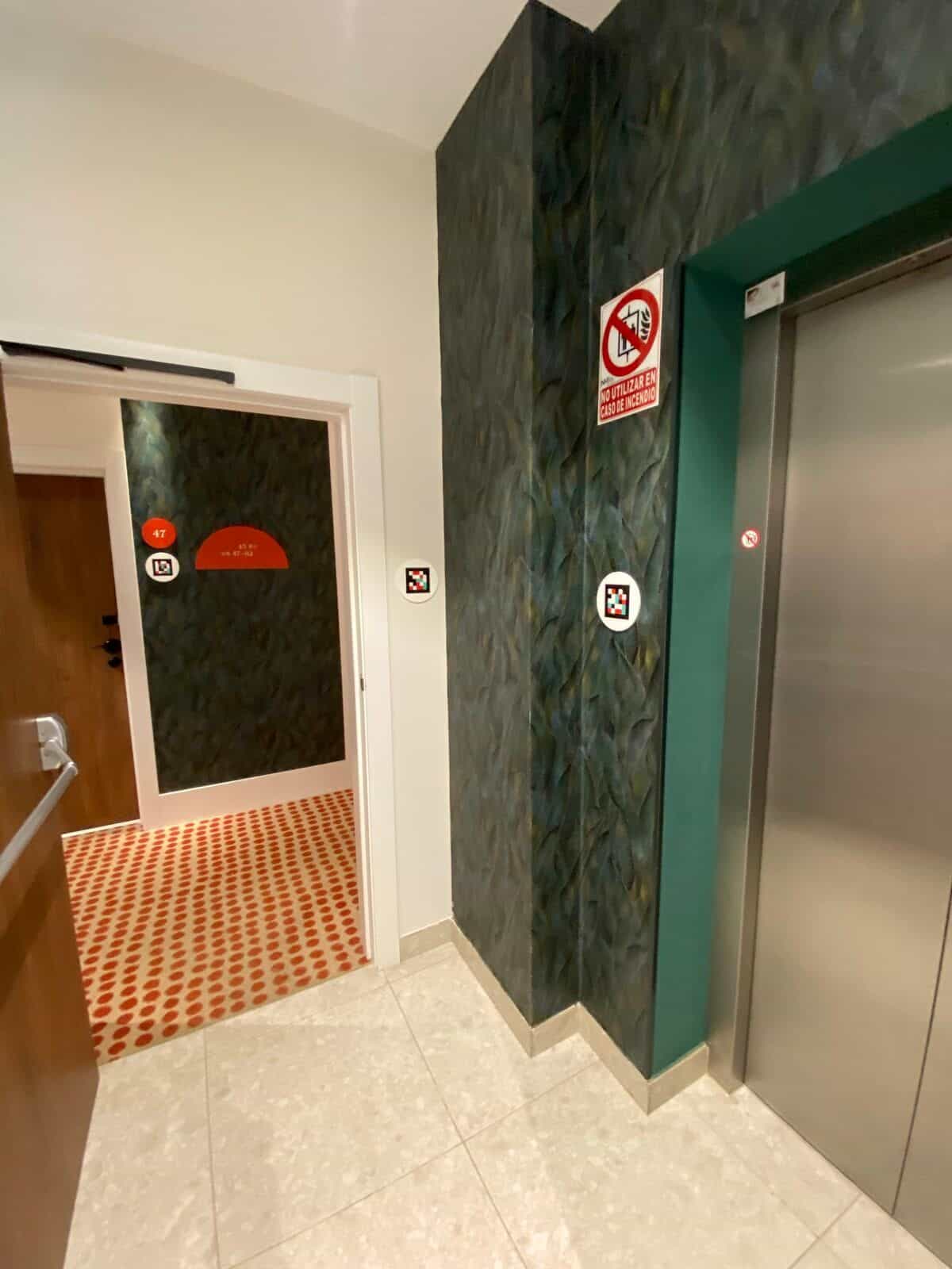 Aufzugsgeschoss am ibis Styles Sevilla: Aufzug mit „im Brandfall nicht benutzen“-Hinweis und NaviLens-Code; weiterer Code an der Frontwand; Flur und Zimmer 47 im Hintergrund