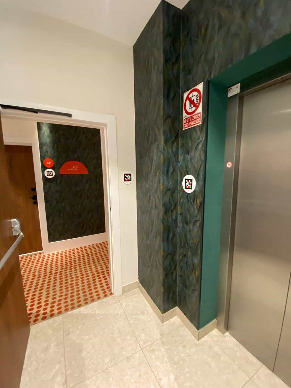 Lift landing at ibis Styles Sevilla: lift with “do not use in case of fire” notice and NaviLens code; another code on the front wall; corridor and Room 47 in the background
