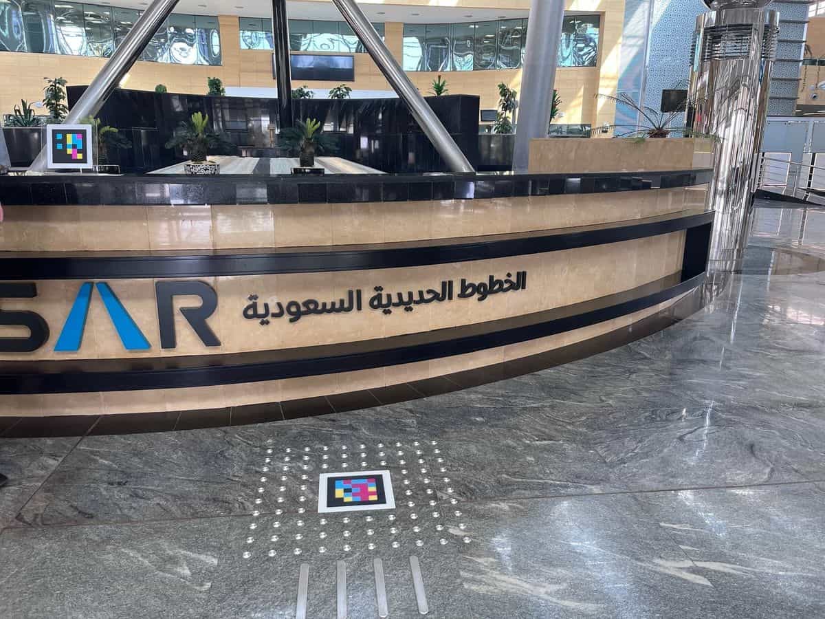 「الخطوط الحديدية السعودية」のロゴが入ったSARインフォメーションカウンター。カウンター上にはスタンド型のNaviLensコード、床には金属製の点字ブロックに囲まれた別のコード