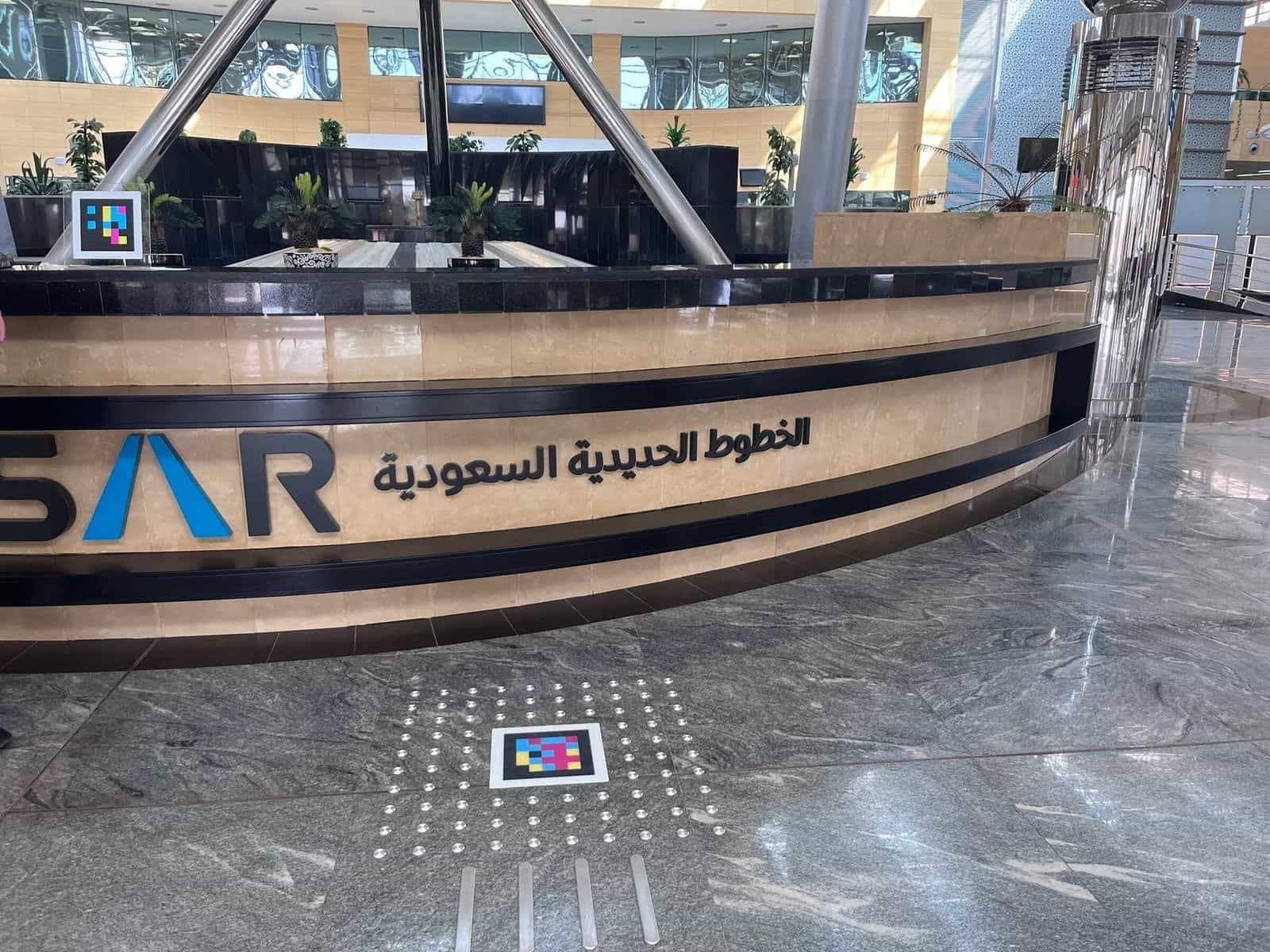 「الخطوط الحديدية السعودية」のロゴが入ったSARインフォメーションカウンター。カウンター上にはスタンド型のNaviLensコード、床には金属製の点字ブロックに囲まれた別のコード