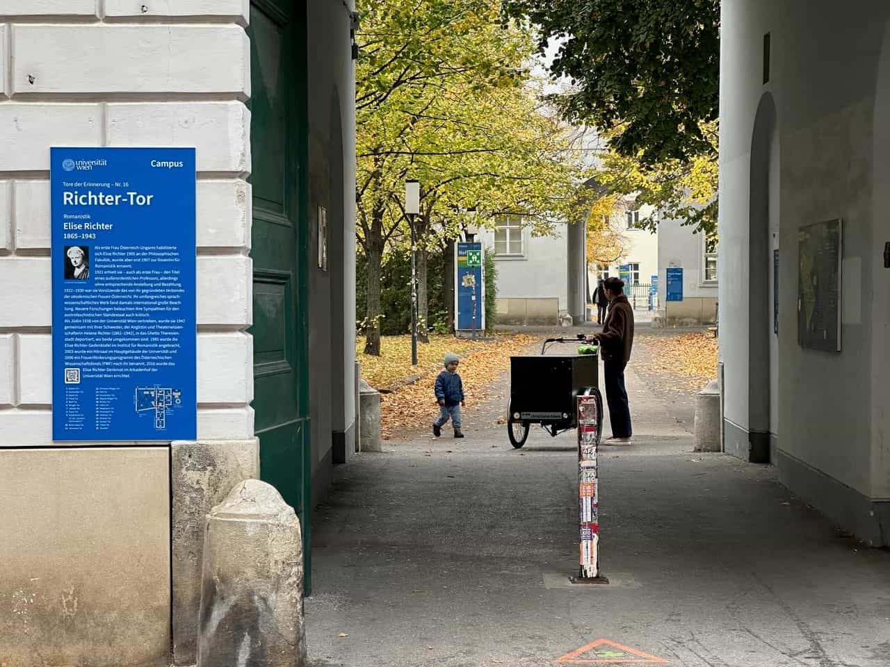 Tafel „Tore der Erinnerung – Nr. 16 Richter-Tor“ für die Romanistin Elise Richter (1865–1943) in einer gewölbten Passage des Campus der Universität Wien; im Hintergrund eine Mutter mit Lastenfahrrad und Kind unter herbstlichen Bäumen