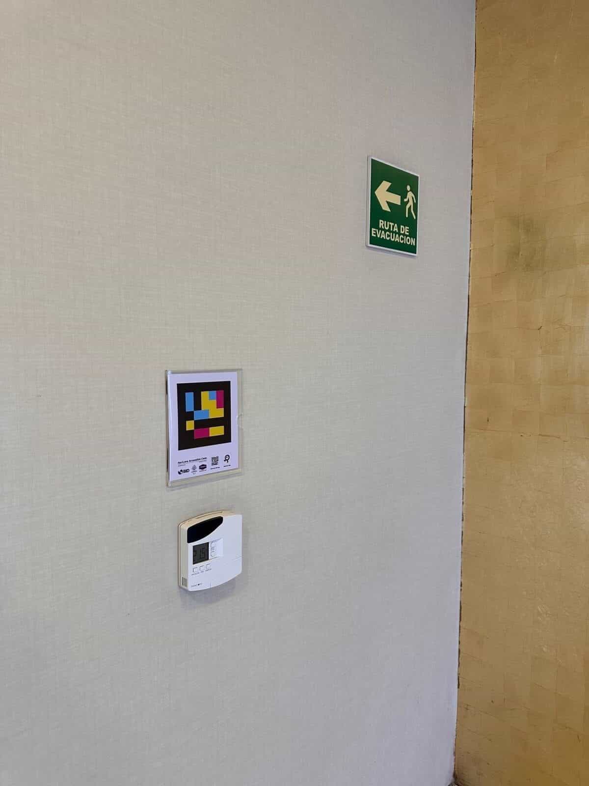Panneau NaviLens sur le mur intérieur à côté du panneau vert « Itinéraire d'évacuation » et du thermostat du couloir