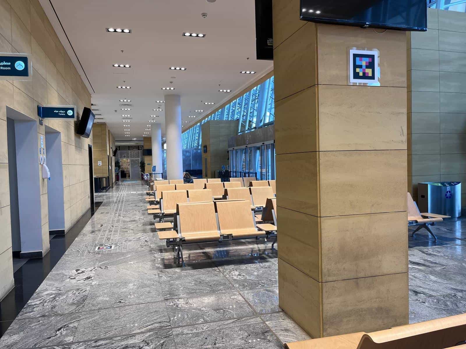 SAR waiting room with wooden seats; on a side column, a NaviLens code with a small “Toilets / دورات مياه” sign pointing to the toilets