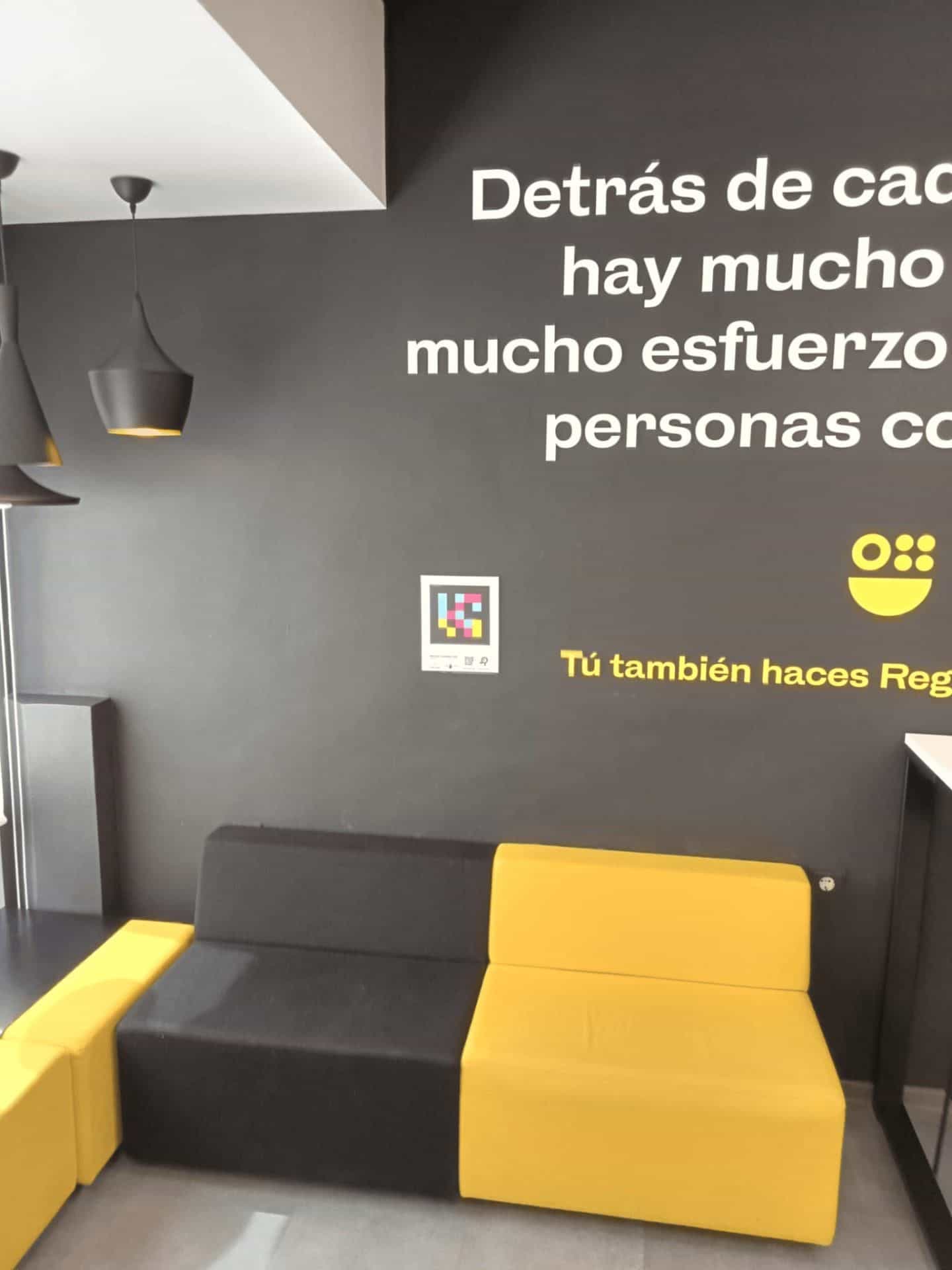 Area di attesa dell’ATRM di Águilas con il murale «Dietro ogni pratica c’è molto lavoro… persone che ci tengono» e il motto giallo «Tú también haces Región», con targa NaviLens accanto al divano modulare nero e giallo.