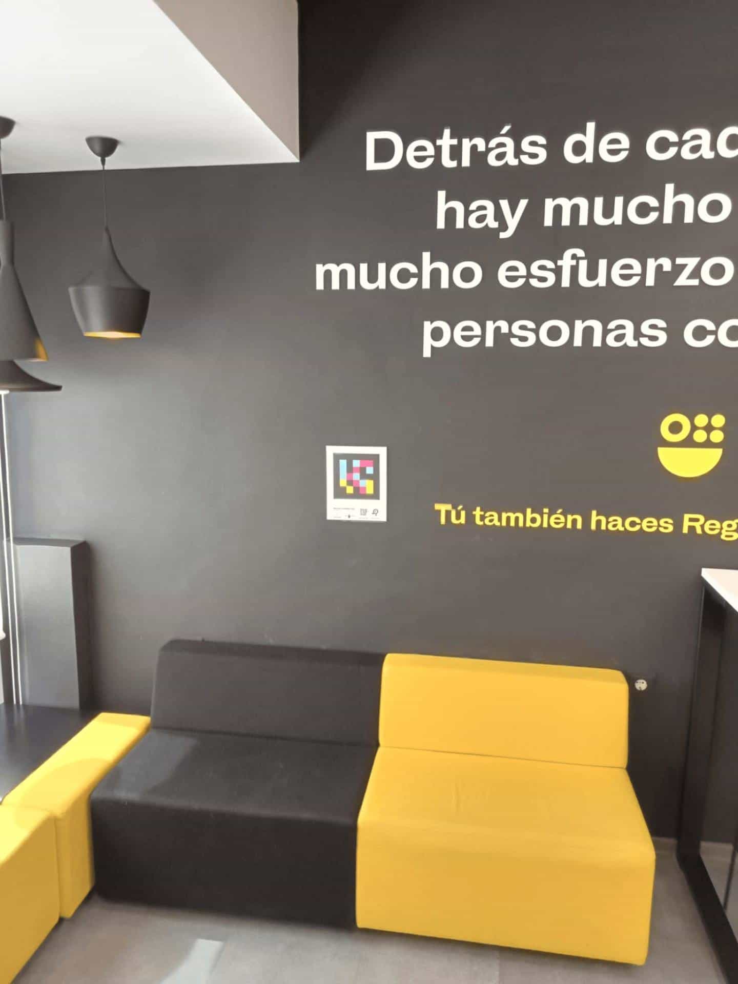 ATRM Águilas waiting area with mural “Behind every step there is hard work… committed people” and yellow motto “You too make Region”, NaviLens plate next to the black & yellow modular sofa.