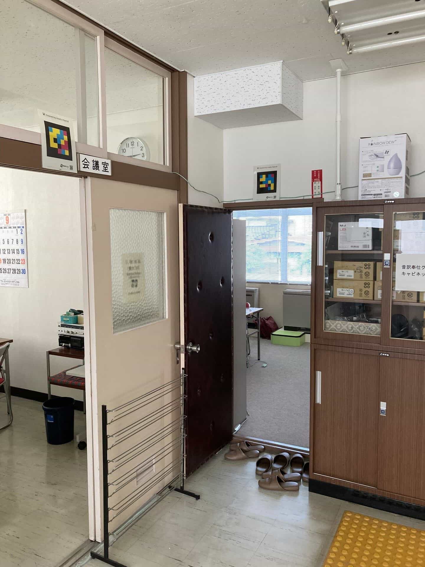 山形県立点字図書館の会議室へのアクセス。ガラス張りの仕切りにある「会議室」の看板の上に1つ、内扉の鴨居の上に1つ、2つのカラフルな四角いNaviLensコードが設置されている。手前には茶色のスリッパと、床には黄色の点字ブロックが見える