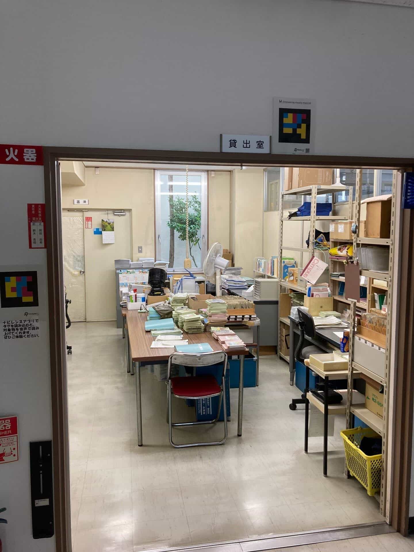 山形県立点字図書館の貸出室の様子。テーブルには点字本が山積みにされ、金属製の棚には読書資料が並んでいる。入口ドアの鴨居の上には、「貸出室」の漢字が書かれた白い看板の隣にカラフルな四角いNaviLensコードがある。側壁には、NaviLensアプリでコードの内容を音声で読み上げられることを説明する案内板が貼られている