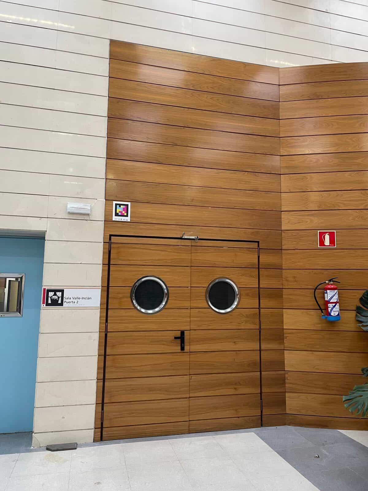Innenraum der RESAD: zweiflügelige Holztür mit zwei runden Metallfenstern, die zum Saal Valle-Inclán führt; an der hellen Marmorwand links der Tür ein weiß gerahmter NaviLens-Code; daneben das Schild „Saal Valle-Inclán · Tür 2“ mit einem roten Vorhangpiktogramm und rechts ein Feuerlöscher
