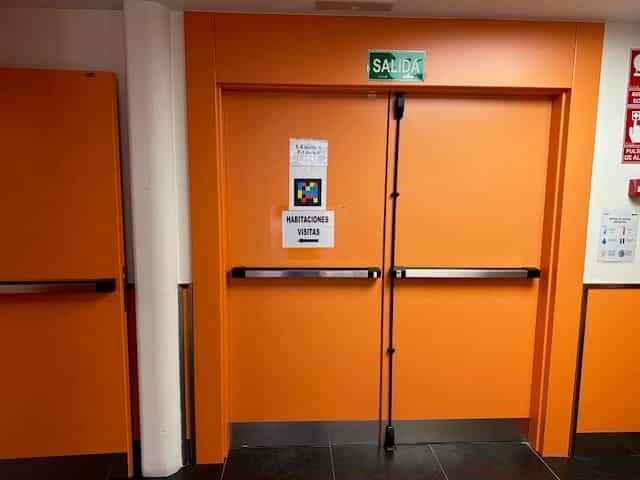 Orange Brandschutztür mit grünem Schild „SALIDA“ (Ausgang), einer Karte mit Pfeil und der Aufschrift „ZIMMER / BESUCH“ und über der Tür ein quadratischer NaviLens-Farbcode im Wohnheimbereich des CRE Madrid