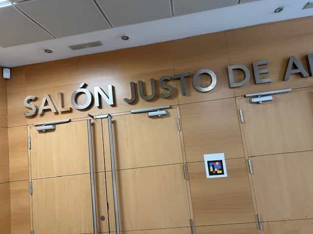Zugang zum „Salón Justo de Aldaco“ im CRE ONCE Madrid: Metallschild mit dem Namen des Saals über Holztüren und daneben eine weiße Plakette mit einem quadratischen NaviLens-Farbcode