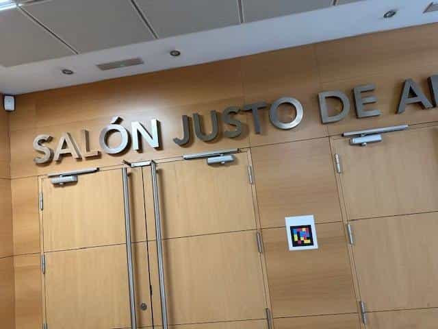 Access to the “Salón Justo de Aldaco” at CRE ONCE Madrid: metal sign with the hall's name above wooden doors and, next to them, a white plate with a square colour NaviLens code