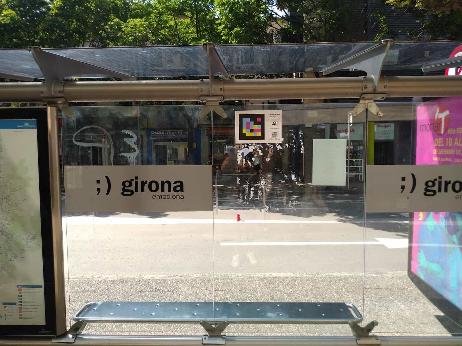 Abribus de Gérone, Carrer de Santa Eugènia · Carrer Illa, avec le panneau « ;) girona emociona », le plan des lignes de la mairie et un code NaviLens sur la vitre