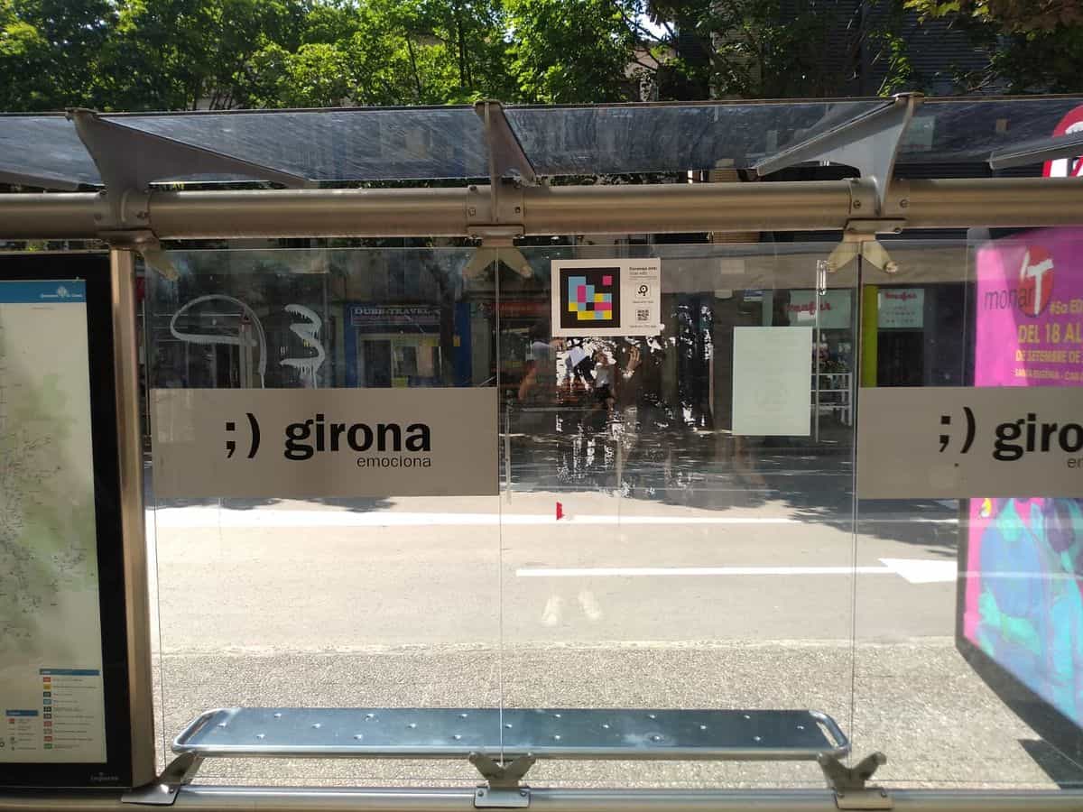 Marquesina del bus de Girona en la calle Santa Eugènia · calle Illa con el cartel «;) girona emociona», el plano de líneas del Ayuntamiento de Girona y un código NaviLens en el cristal