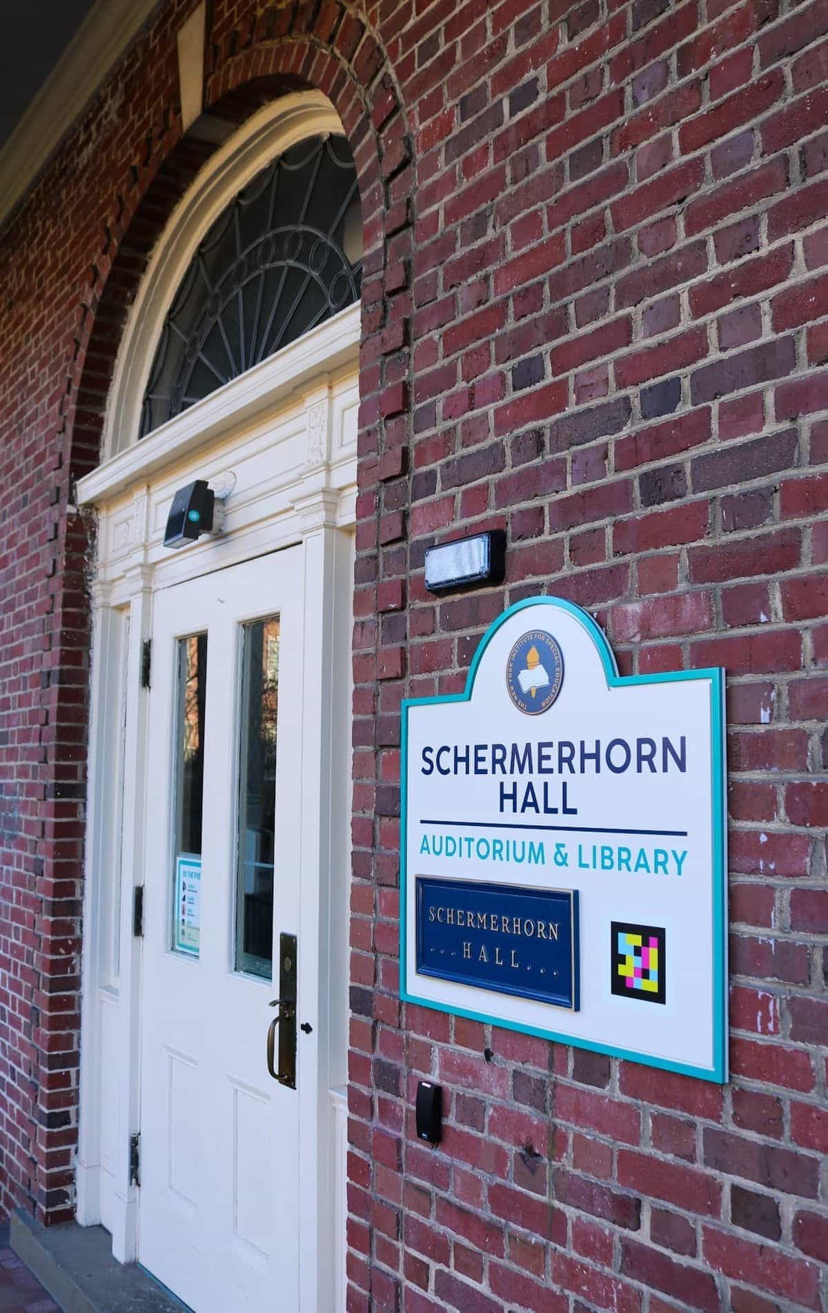 Entrance to NYISE's Schermerhorn Hall: white arched door on red-brick facade with a turquoise-framed sign reading 'SCHERMERHORN HALL · Auditorium & Library' and a NaviLens code that reads the entrance