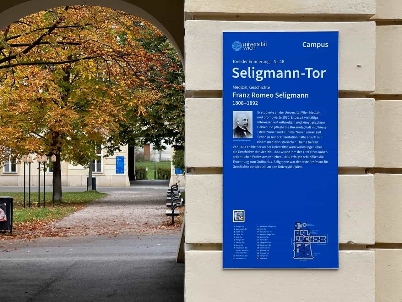 Blaue Tafel „Tore der Erinnerung – Nr. 18 Seligmann-Tor“ für Franz Romeo Seligmann (1808–1892) in einer Passage des Campus der Universität Wien; am Tafelfuß NaviLens-Code, Liste der 23 Tore und schematischer Campusplan