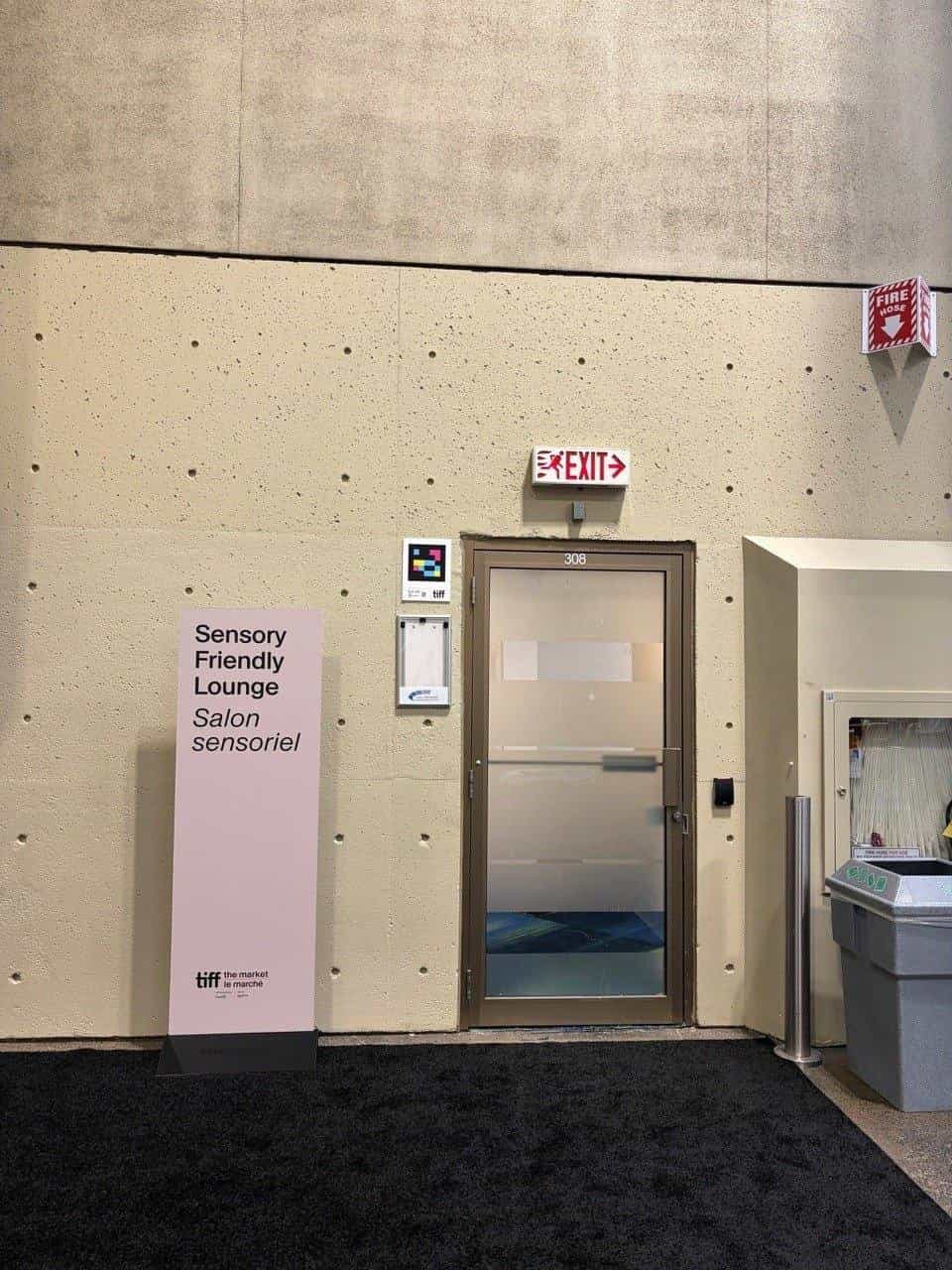The door of room 308 at the Metro Toronto Convention Centre, with a pink sign reading “Sensory Friendly Lounge · Salon sensoriel” under the tiff · the market logo. On the concrete wall, to the left of the door, a NaviLens code.