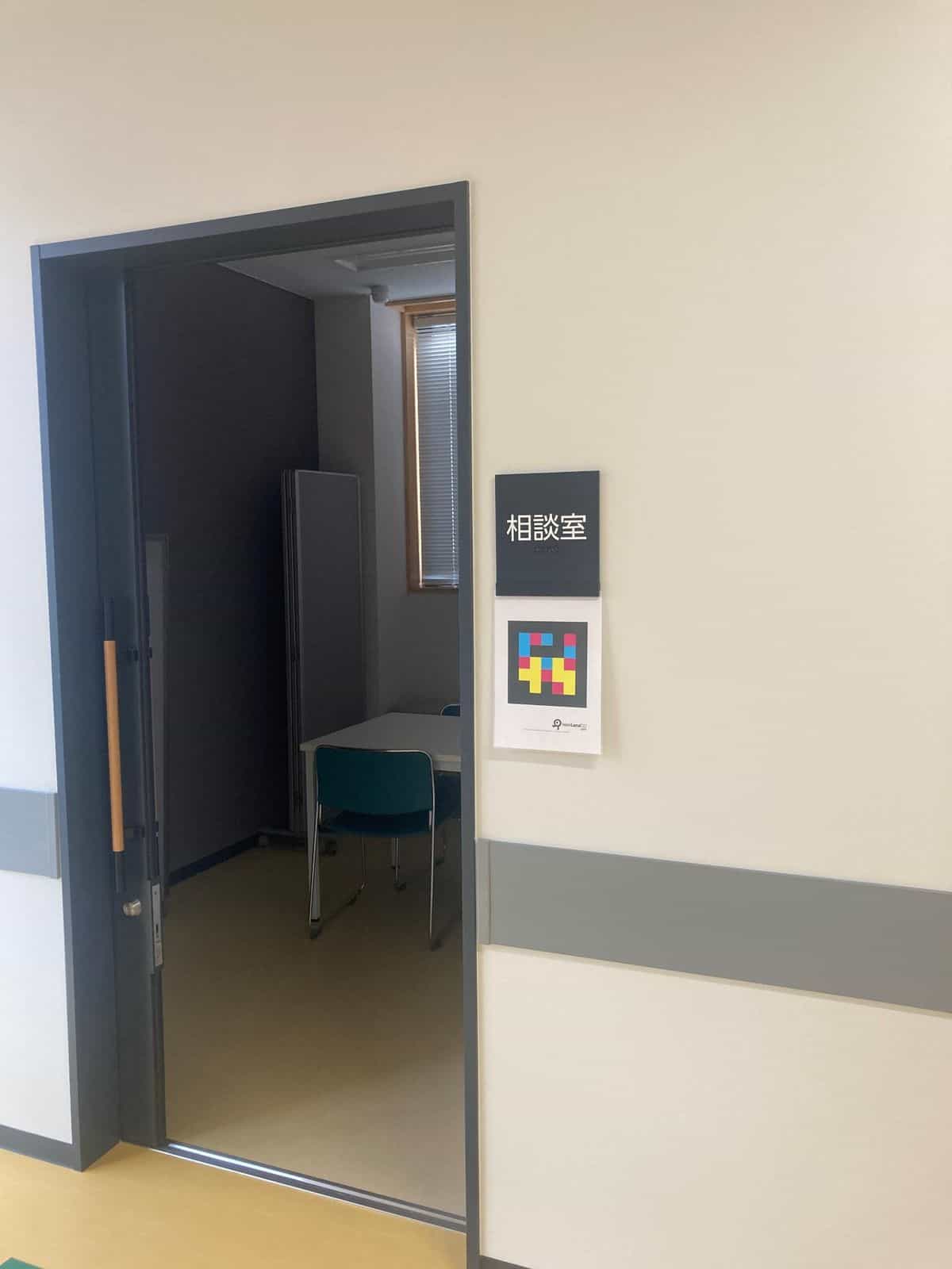 Puerta entreabierta de la sala de consulta («相談室»): se ven en el interior una mesa pequeña gris con sillas verde-azuladas y un panel acústico oscuro plegado contra la pared, junto a una ventana con persiana veneciana; a la derecha de la puerta, placa táctil negra con texto blanco «相談室» y braille en la zona inferior, y debajo un cartel NaviLens GO con código en colores cian, magenta y amarillo sobre fondo negro; rodapié gris a media altura