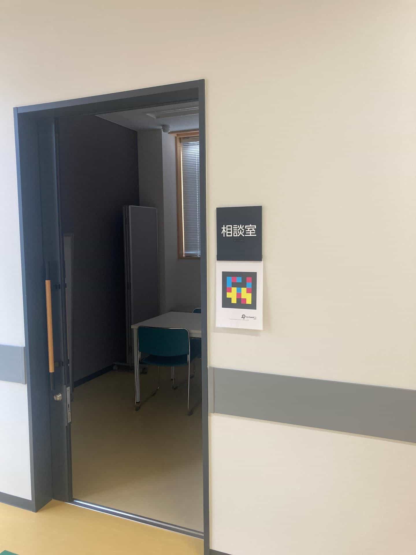 Porta socchiusa della sala consulenza («相談室»): all’interno si vedono un piccolo tavolo grigio con sedie verde-azzurre e un pannello acustico scuro ripiegato contro la parete, accanto a una finestra con veneziana; a destra della porta, targa tattile nera con il testo bianco «相談室» e braille nella parte inferiore, e sotto un cartello NaviLens GO con il codice in ciano, magenta e giallo su fondo nero; zoccolino grigio a mezza altezza