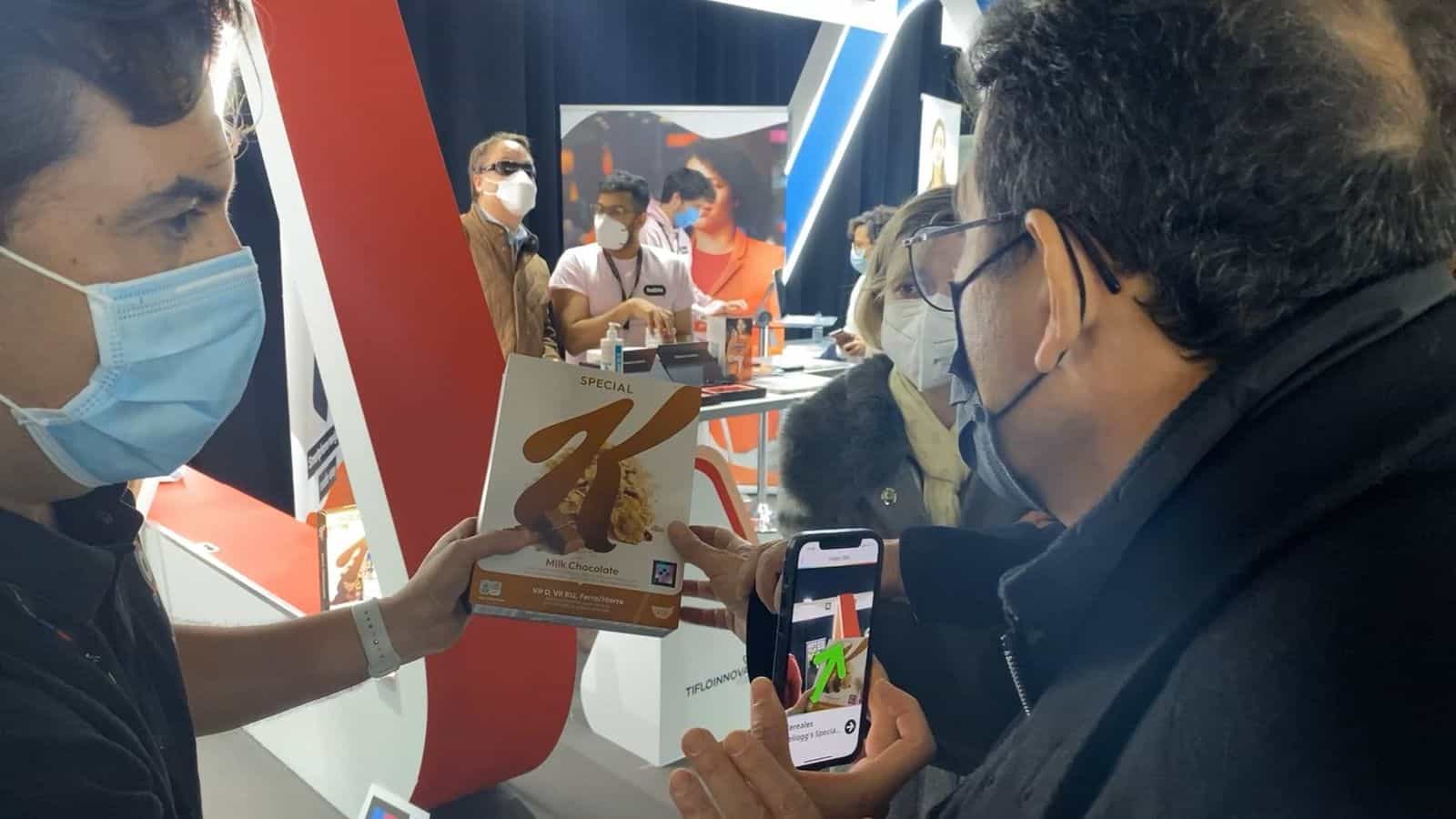 Un visitatore di Tifloinnova 2022 tiene il suo iPhone per inquadrare il codice NaviLens stampato su una confezione Special K «Milk Chocolate» che un tecnico di NaviLens, con mascherina chirurgica azzurra, gli avvicina; lo schermo del telefono mostra una freccia verde che indica dove si trova il codice e, in fondo, altri visitatori con bastoni bianchi provano le demo nello stand
