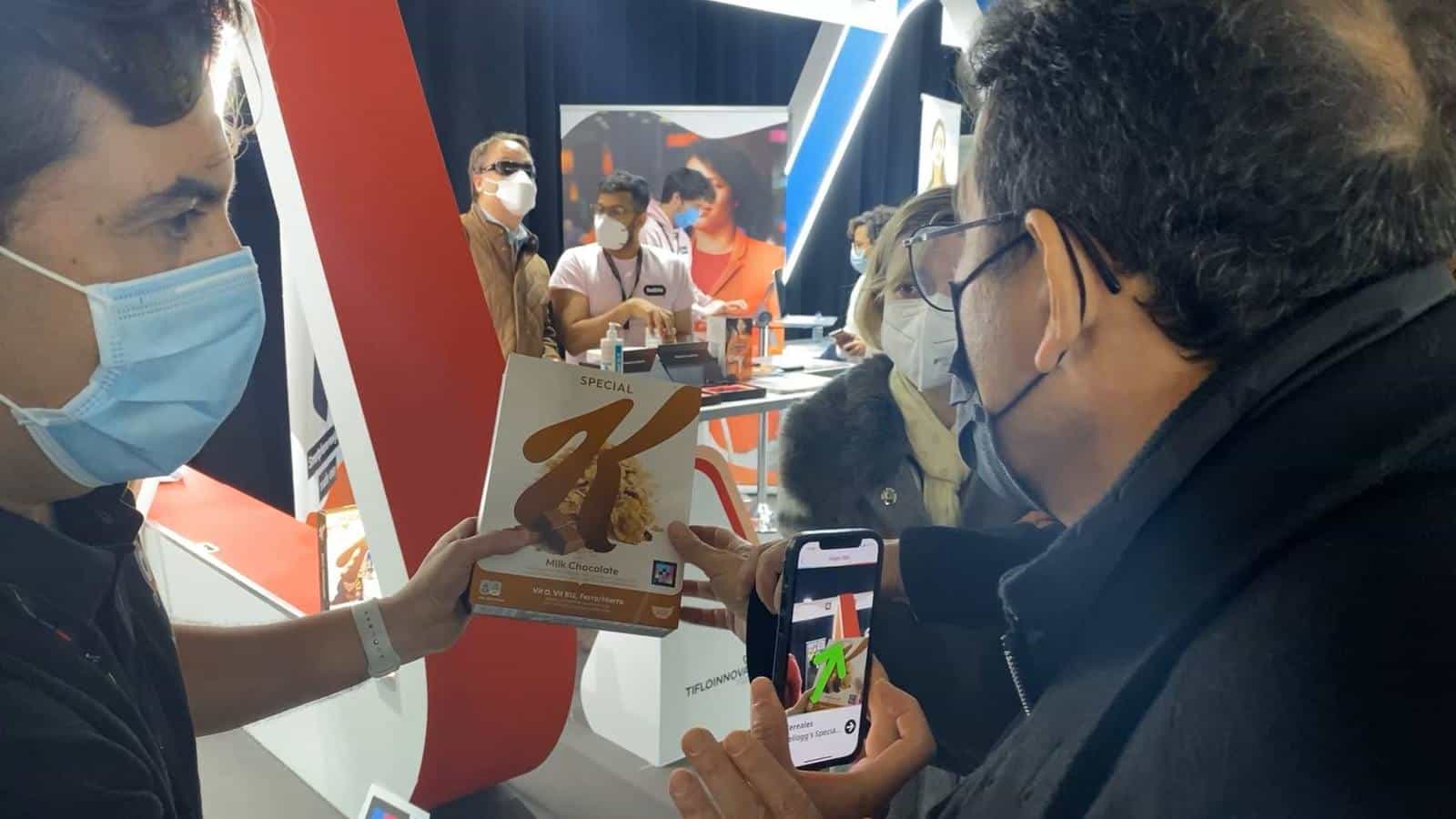 A visitor at Tifloinnova 2022 holds up his iPhone to scan the NaviLens code printed on a Special K “Milk Chocolate” box that a NaviLens technician in a blue surgical mask is handing him; the phone screen shows a green arrow indicating where the code is and, in the background, other visitors with white canes try out demos at the stand