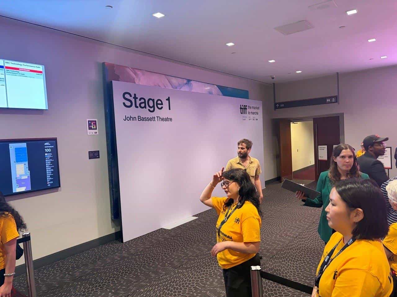 The entrance to Stage 1, the John Bassett Theatre, during TIFF: The Market. On the wall to the left, a NaviLens code next to the screens showing the day's programme; in front, festival volunteers in yellow T-shirts direct delegates towards the theatre.