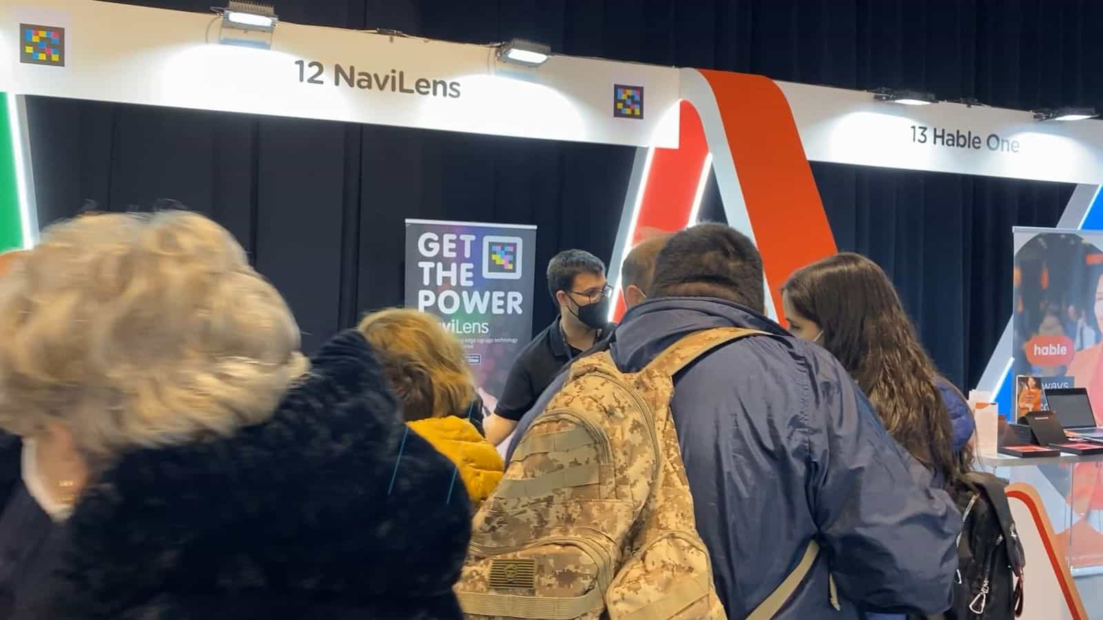 Testata dello stand 12 «NaviLens» a Tifloinnova 2022, con un codice NaviLens accanto all’insegna e un altro accanto allo stand 13 «Hable One»; un gruppo di visitatori —fra cui una persona con giacca a vento mimetica e un’altra con anorak giallo— si accalca davanti a un tecnico di NaviLens con polo nero e mascherina, accanto al rollup «GET THE POWER · NaviLens»