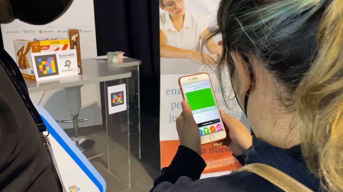 Una visitante de Tifloinnova escanea con su iPhone un código NaviLens colocado sobre una peana en el stand 12; junto al código, un cartel «Scan with NaviLens GO» y dos cajas de cereales Special K Classic con código NaviLens y precio 1,99 €. La pantalla del móvil muestra el resultado del escaneo en una franja verde con el texto «Stop NaviLens»