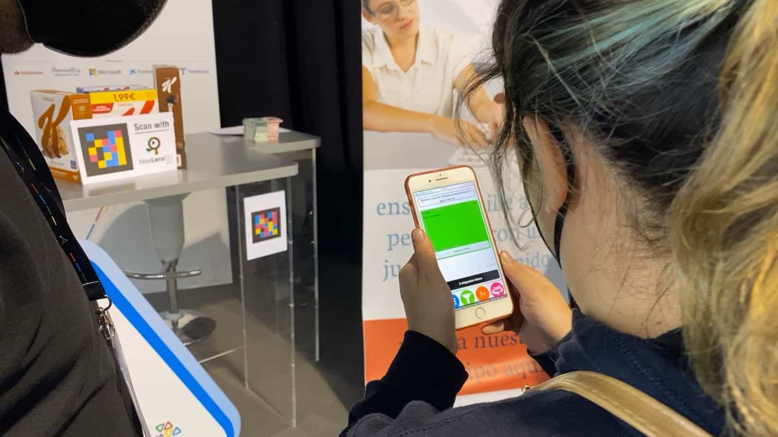 Una visitatrice di Tifloinnova inquadra con il suo iPhone un codice NaviLens collocato su un basamento nello stand 12; accanto al codice, un cartello «Scan with NaviLens GO» e due confezioni di cereali Special K Classic con codice NaviLens e prezzo 1,99 €. Lo schermo del telefono mostra il risultato della scansione in una fascia verde con il testo «Stop NaviLens»